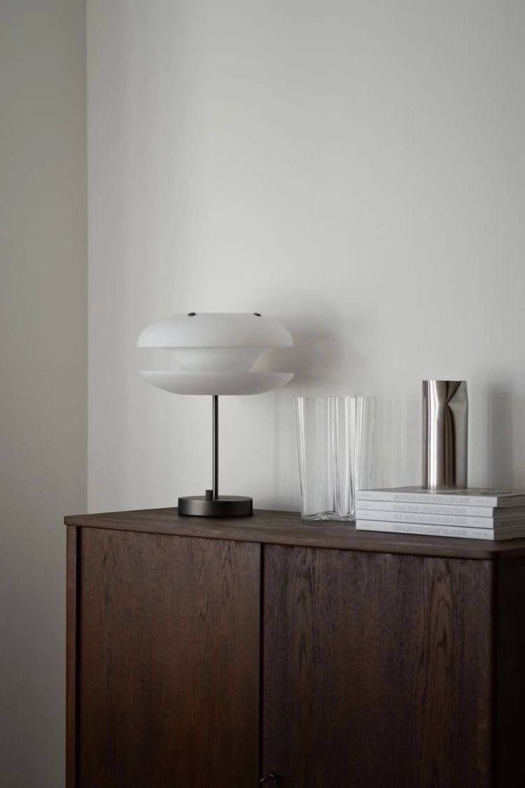 A styled interior scene featuring a chocolate-coloured chest of drawers topped with a white sculptural lamp, a glass jug of water, stacked books, and a small silver decorative object, arranged in a warm, minimal, and earthy aesthetic.